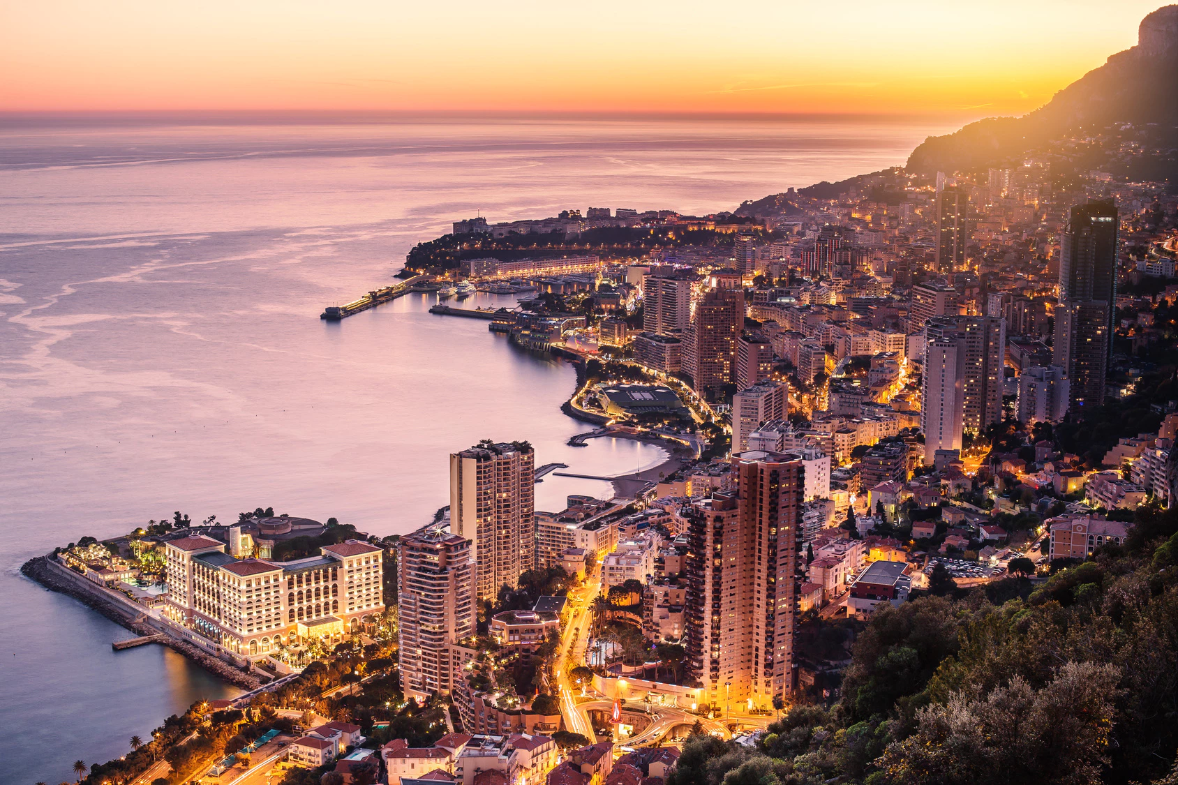 Aerial view of Monaco's glowing city lights and coastline during a vibrant Mediterranean sunset.